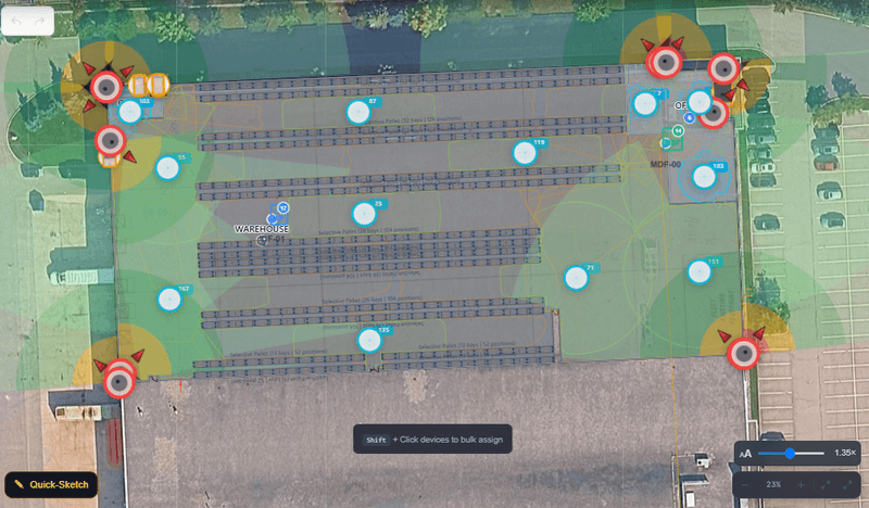 A warehouse floor plan anchored over satellite imagery of the real building, with the racking aisles and pallet-position labels drawn to scale. Twelve wireless access points sit as blue markers through the aisles, and perimeter cameras show shaded coverage wedges reaching across the yard and loading area.